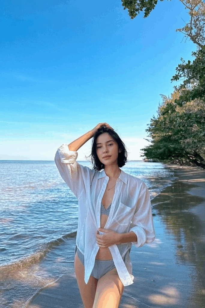 wearing an oversized, sheer white cotton button-down shirt, sleeves rolled, tied loosely at the waist over a striped bikini