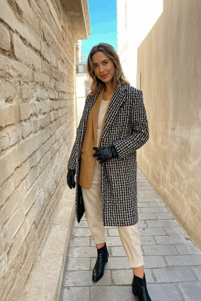 A woman wearing a textured tweed coat in shades of black and cream over a monochrome tan ensemble. She finishes the look with black leather gloves and low-heeled boot