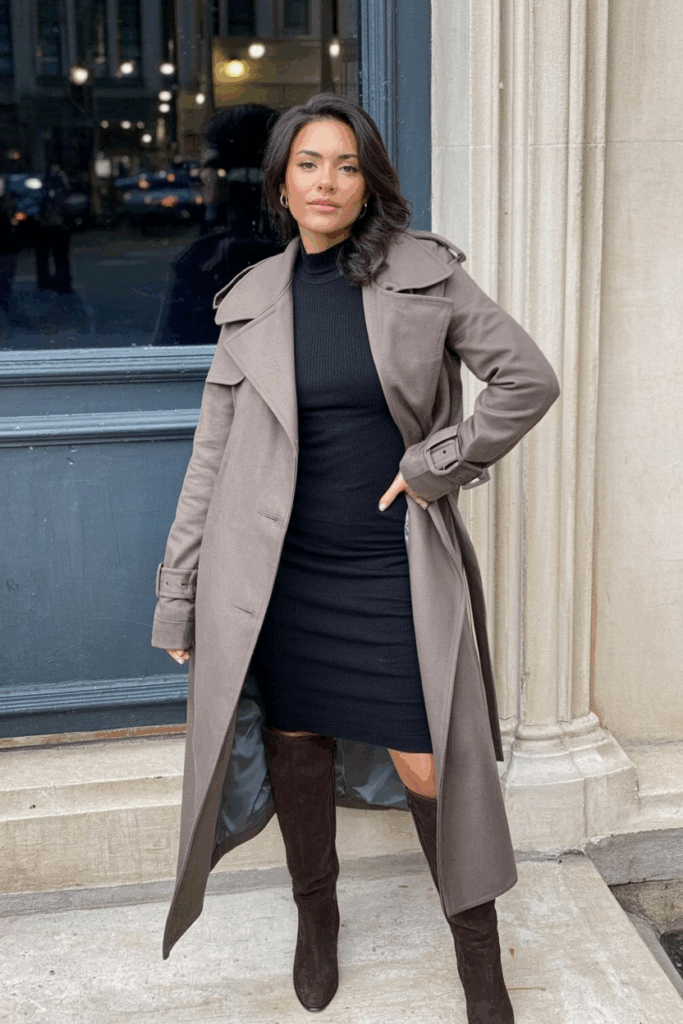 A long taupe trench-style coat in thick wool with storm flaps and a matching belt, worn over a fitted knit dress and tall suede boots