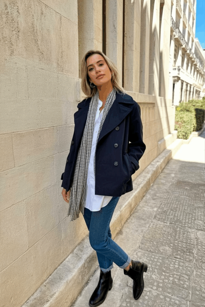A woman wearing a navy peacoat, checked scarf, jeans, and ankle boots