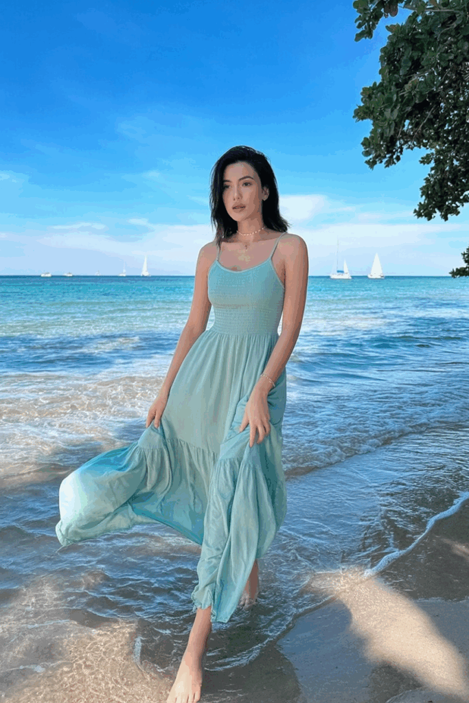 A woman in a flowing seafoam green spaghetti-strap maxi dress, standing barefoot in the shallow edge of the ocean, dress swaying at ankle length