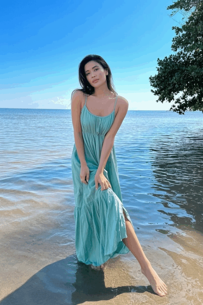 A woman in a flowing seafoam green spaghetti-strap maxi dress, standing barefoot in the shallow edge of the ocean, dress swaying at ankle length, gaze soft