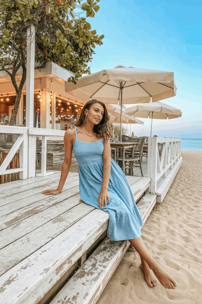 A woman in a soft pastel blue cotton sundress with thin straps and a flowing skirt, standing near a beachside café