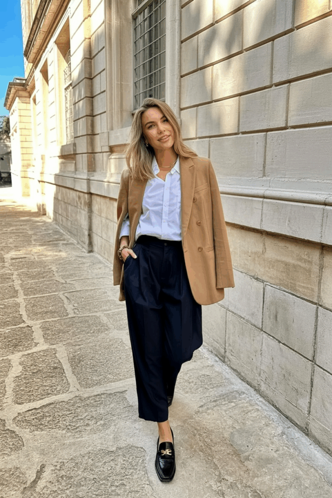 A woman wearing a camel wool blazer over a white button-up shirt and navy pleated trousers with black leather loafers