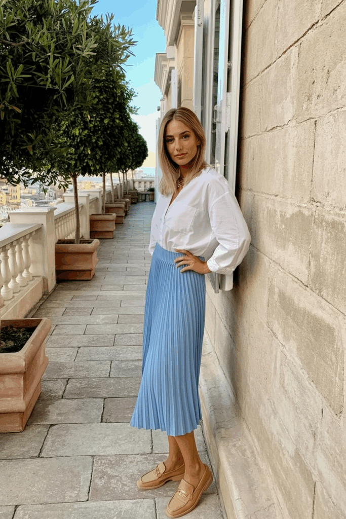 A woman wearing a white blouse tucked into a powder blue pleated skirt with tan loafers