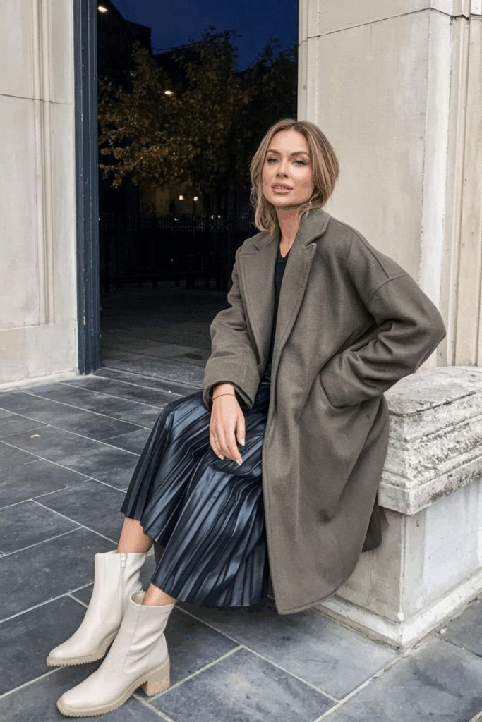 A woman wearing an oversized alpaca-blend coat in muted olive over a pleated skirt and cream ankle boots
