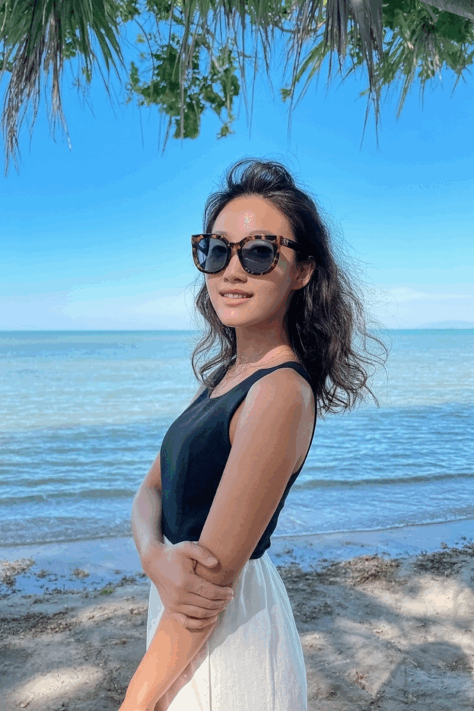 classic tortoiseshell oversized sunglasses, smiling softly, with the ocean blurred in the background.