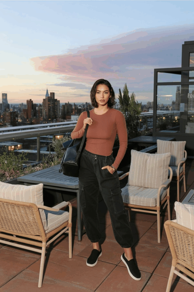 soft black cargo pants, a long-sleeve ribbed rust-colored crewneck, and black slip-on sneakers, carrying a small backpack.
