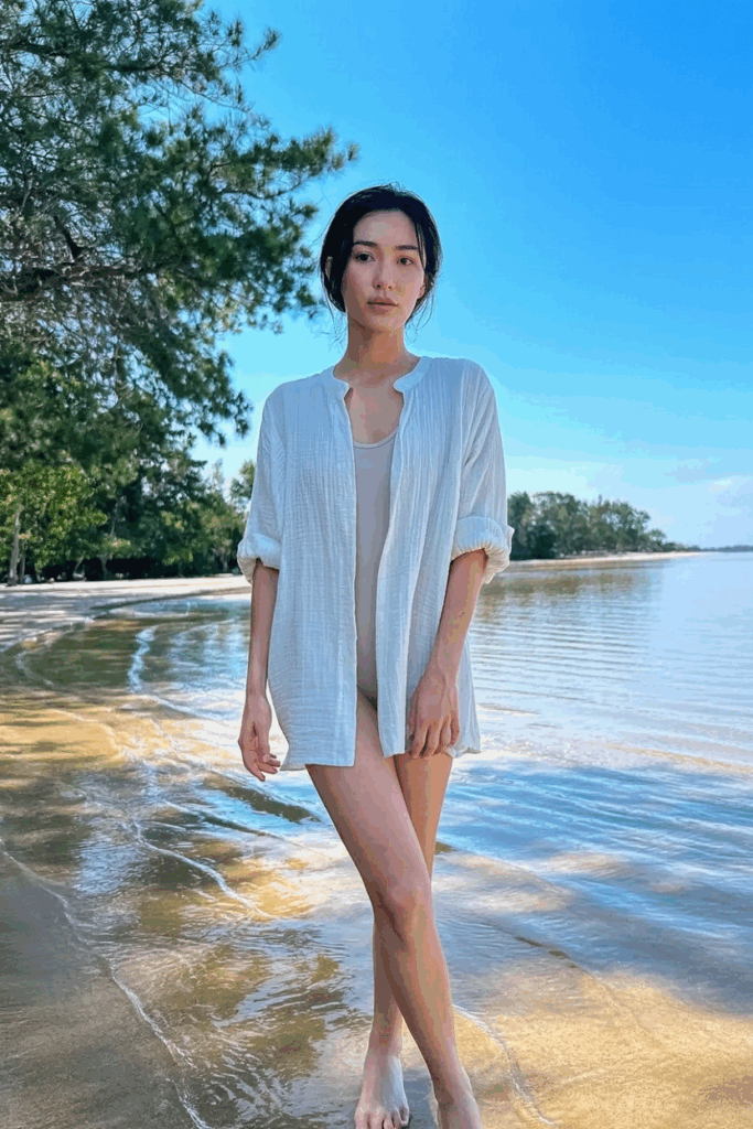 wearing a loose, knee-length white linen cover-up with rolled sleeves, paired with a simple beige swimsuit underneath, walking barefoot on sand.