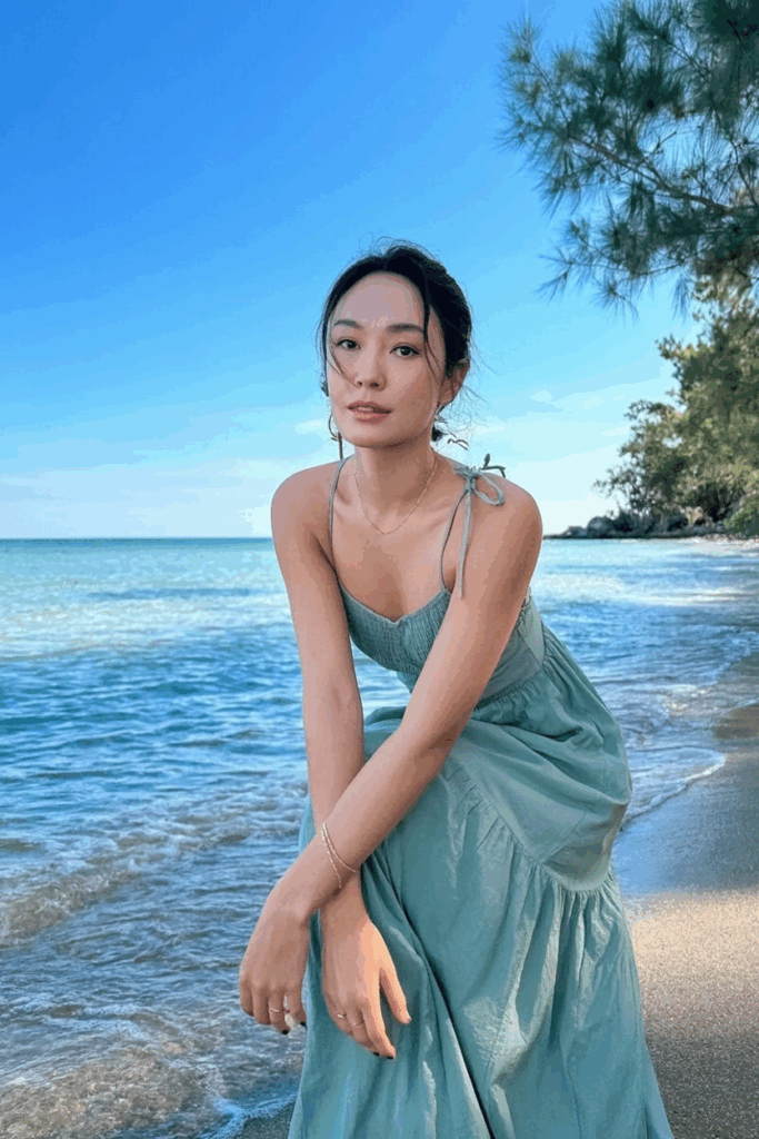 wearing a seafoam green flowing maxi dress with thin straps, minimal gold jewelry, standing near turquoise ocean water.