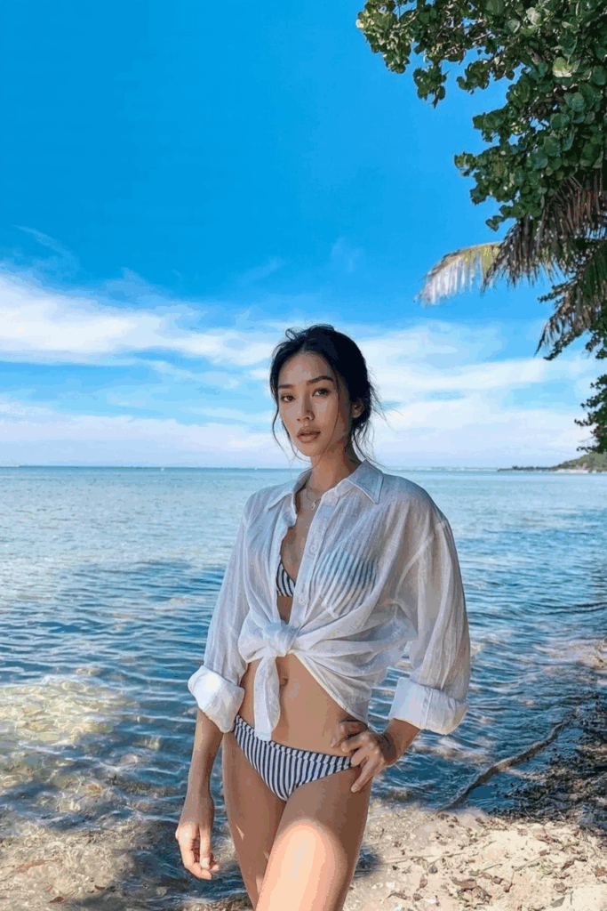 wearing an oversized, sheer white cotton button-down shirt, sleeves rolled, tied loosely at the waist over a striped bikini.