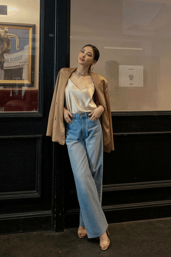 wearing light blue wide-leg jeans, a cream silk camisole, a tan blazer draped over her shoulders, and nude heels