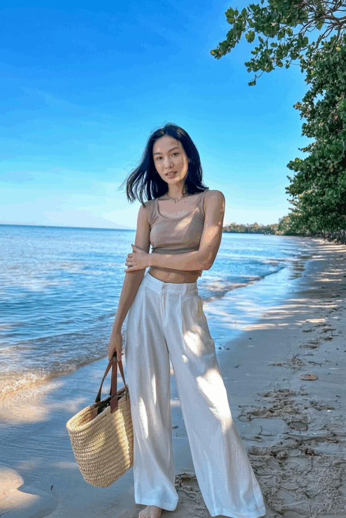 wearing white wide-leg linen pants paired with a tucked-in beige tank top, barefoot on a sandy beach, carrying a straw tote bag.