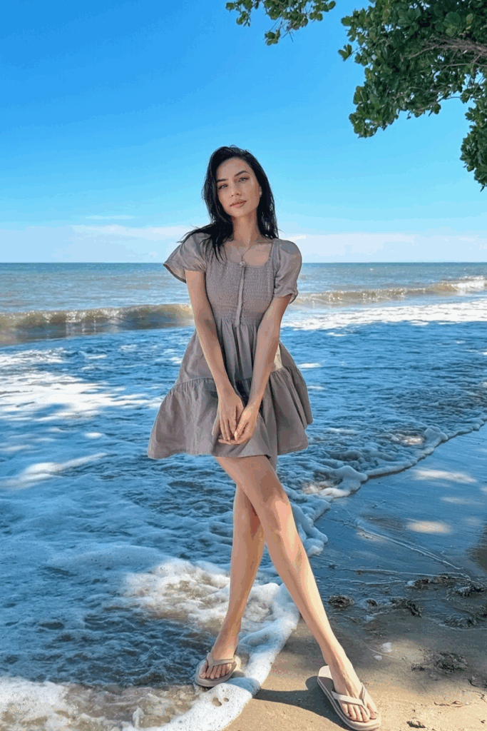 woman in a short airy taupe dress with neutral flip-flops, long legs visible, standing where waves meet the shore
