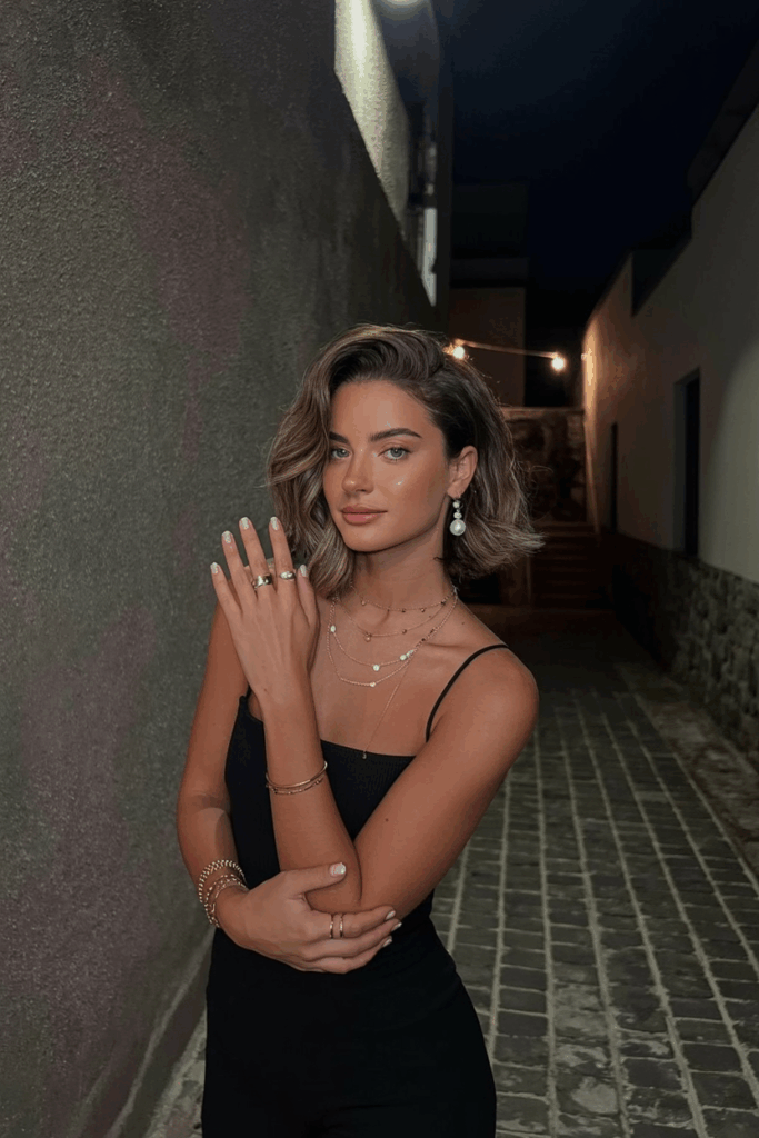 woman wearing layered gold necklaces, thin stacking rings, and minimal pearl drop earrings. Her nails are short and polished in nude